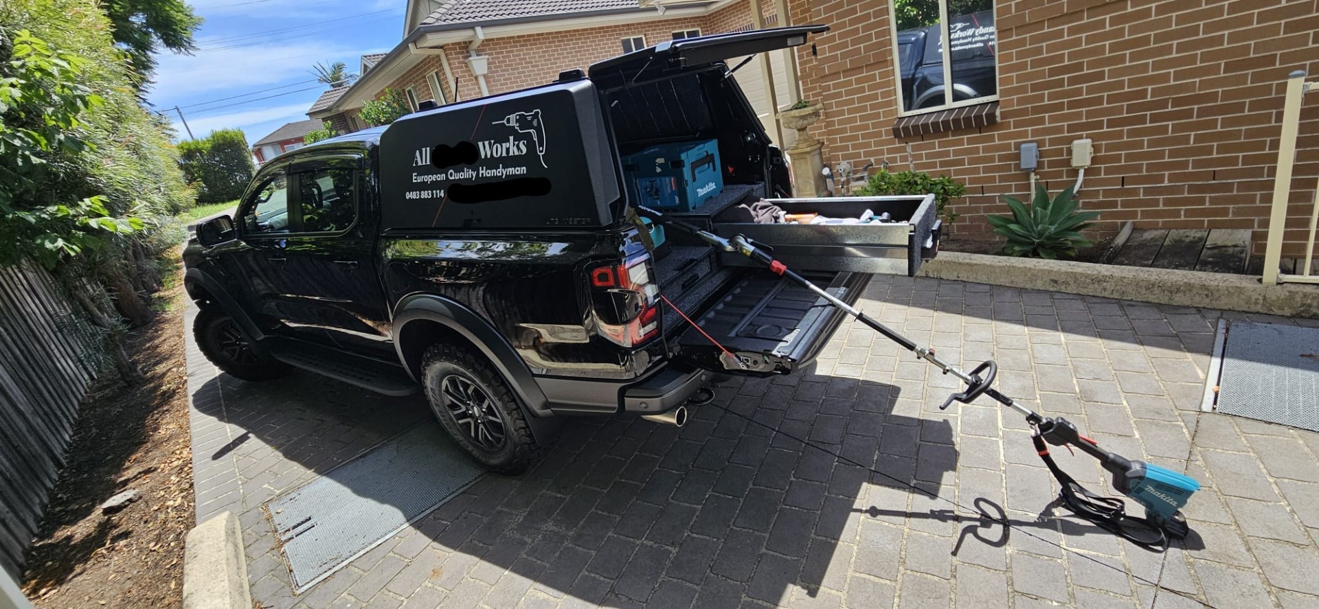 Black service truck parked in driveway with open tailgate and tools, including a blue pressure washer.
