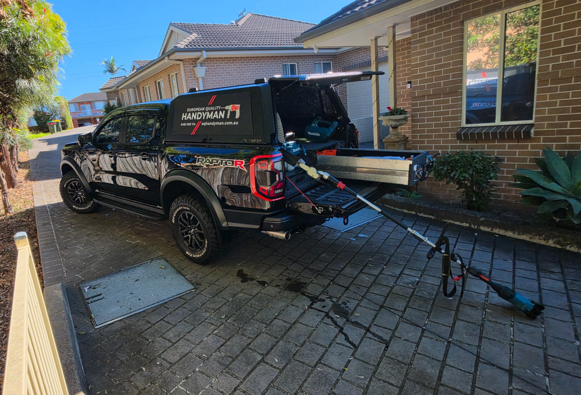Black service truck parked in driveway with open tailgate and tools, including a blue pressure washer.